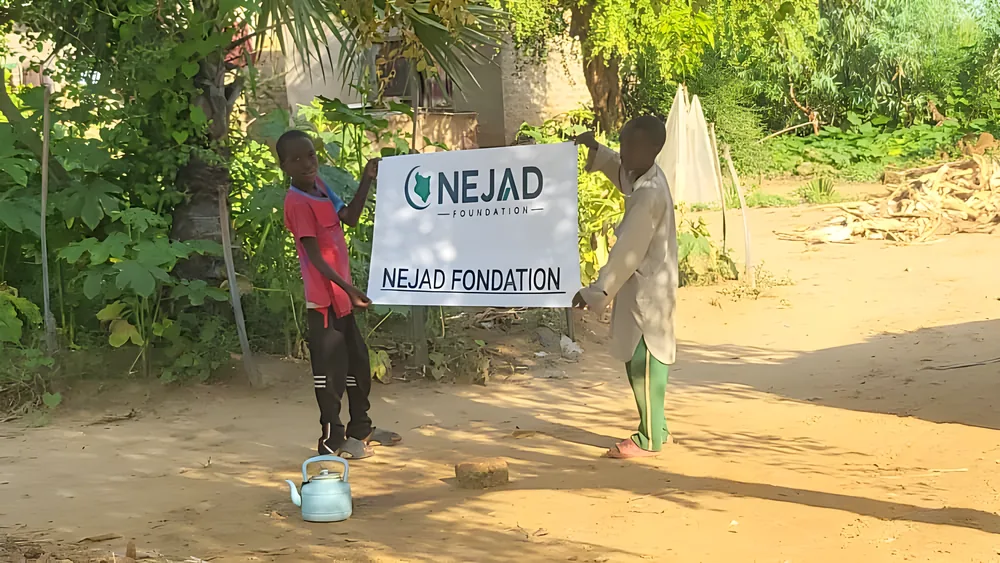 Two children hold a NEJAD Foundation banner in a sandy yard, a water kettle at their feet
