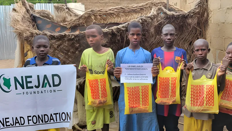 Children holding 'Thank you' bags of Qurbani meat beside a NEJAD Foundation banner
