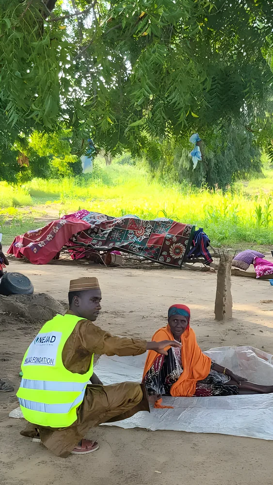 A NEJAD volunteer kneels to speak with an elderly woman sitting on a mat outdoors