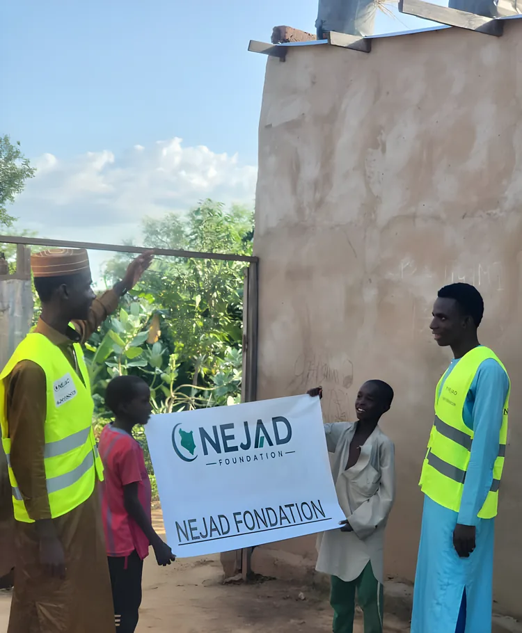 A NEJAD volunteer points to the NEJAD banner held up by two boys