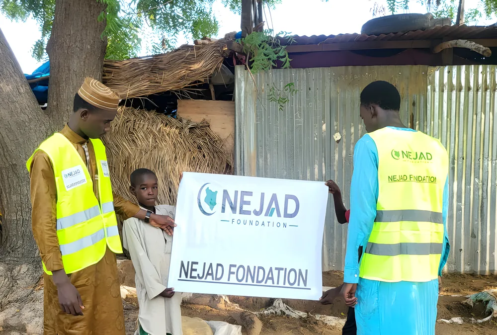 Two NEJAD volunteers and a boy holding the NEJAD banner outside a home