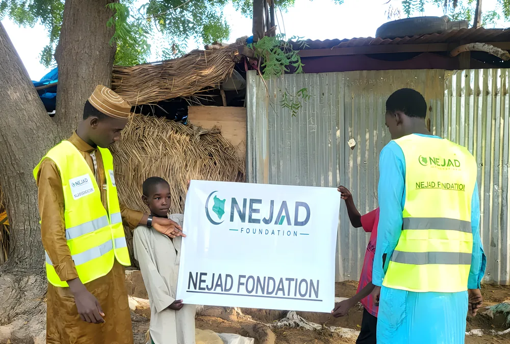 NEJAD Foundation volunteers in high-visibility vests holding the NEJAD banner with a young boy